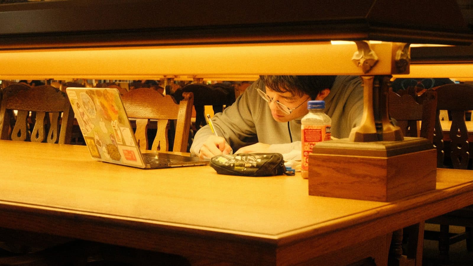 A student writes by hand under a library lamp, with a laptop pushed to one side of the desk.