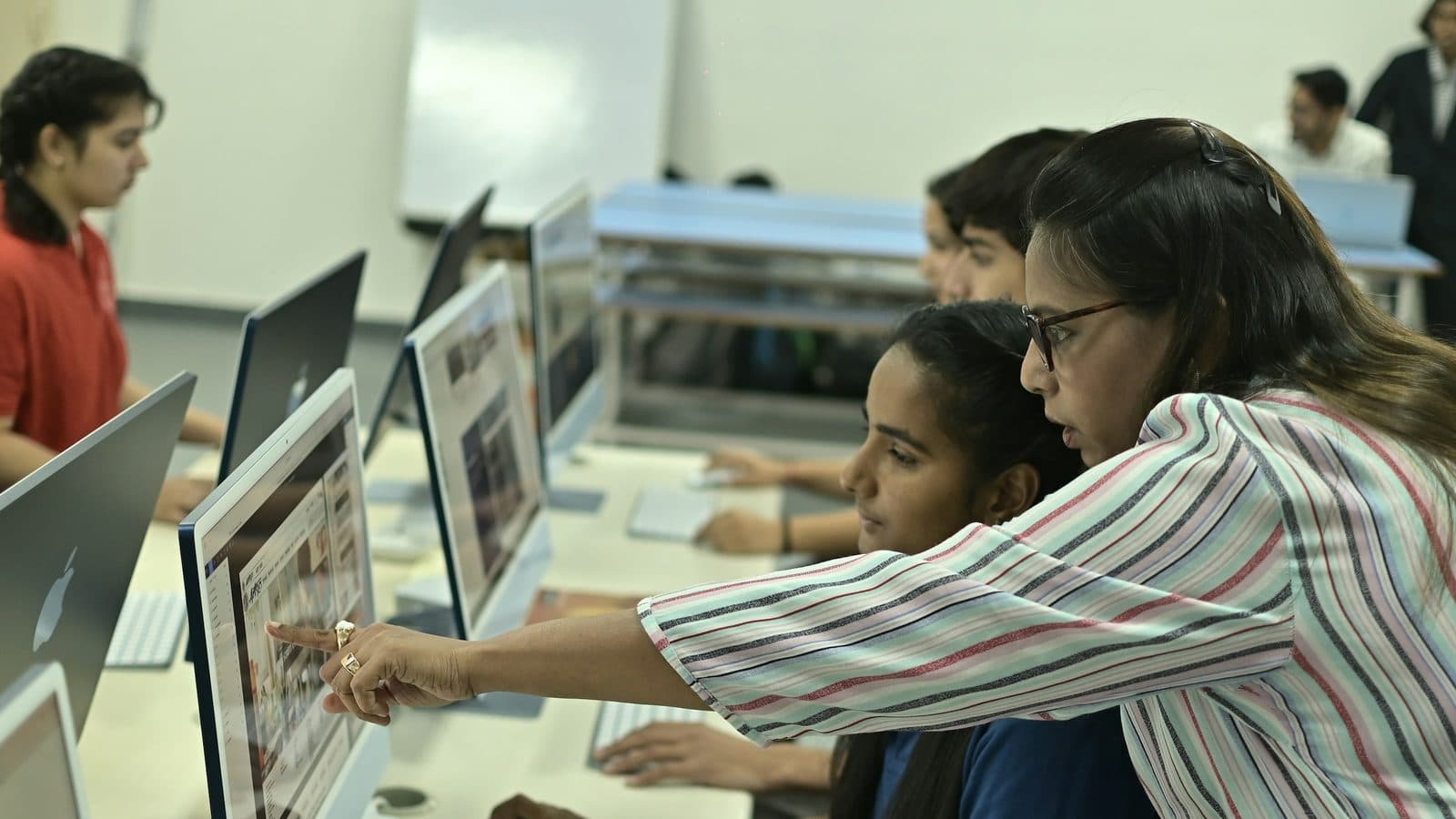 A teacher points at a computer screen as she guides a student in a school computer lab.
